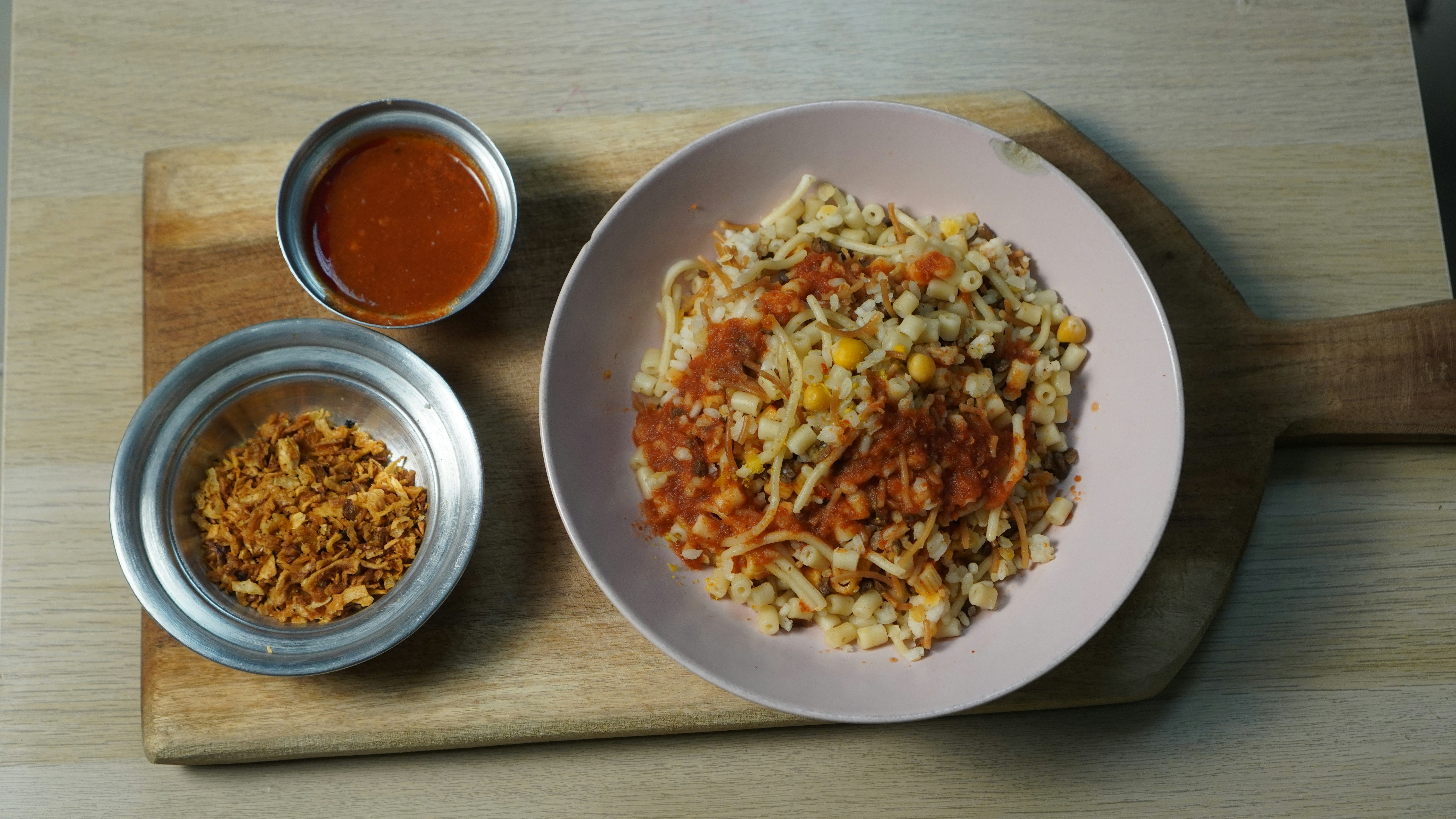A small bowl of koshari, with a cup of sauce and a katori of crispy fried onions on its side.